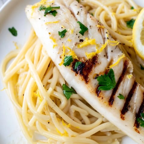 Grilled lemon bass fillets rest atop spaghetti tossed in a zesty garlic butter sauce with fresh parsley garnish.