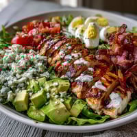 Freshly grilled chicken slices and crispy bacon over a vibrant Cobb Salad Bowl, topped with creamy avocado and tangy blue cheese crumbles.
